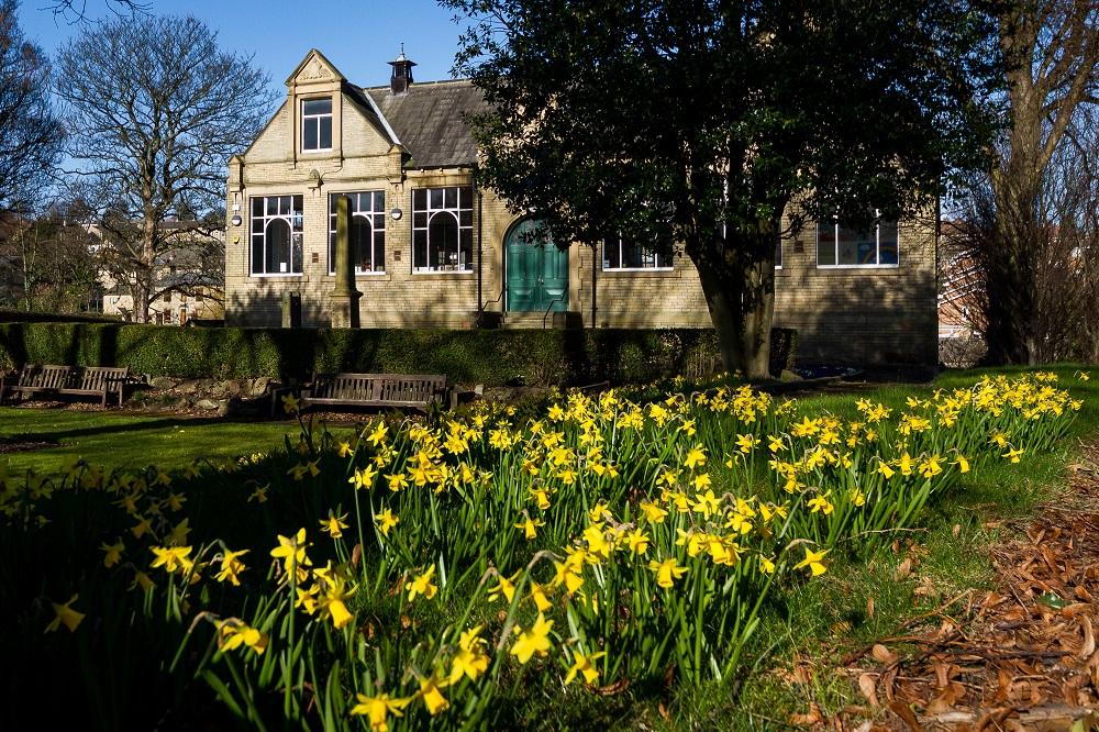 Rastrick Library in Spring. The Library is in the background, against a blue sky. A flowerbed of daffodils is in the foreground.